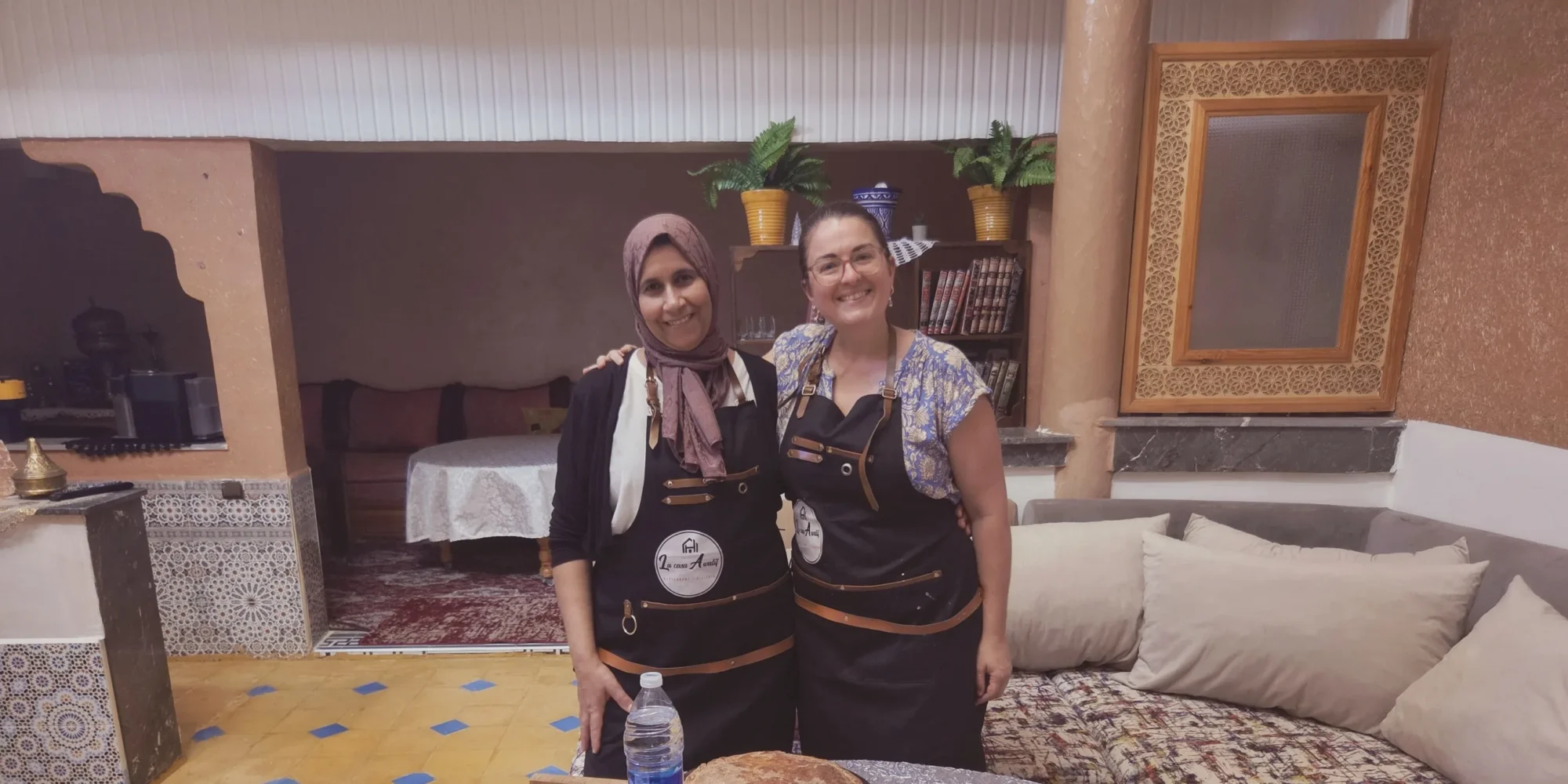 Cours de cuisine à Ouarzazate : Aouatif et son invitée, souriantes en tablier, dans le salon marocain de la maison.