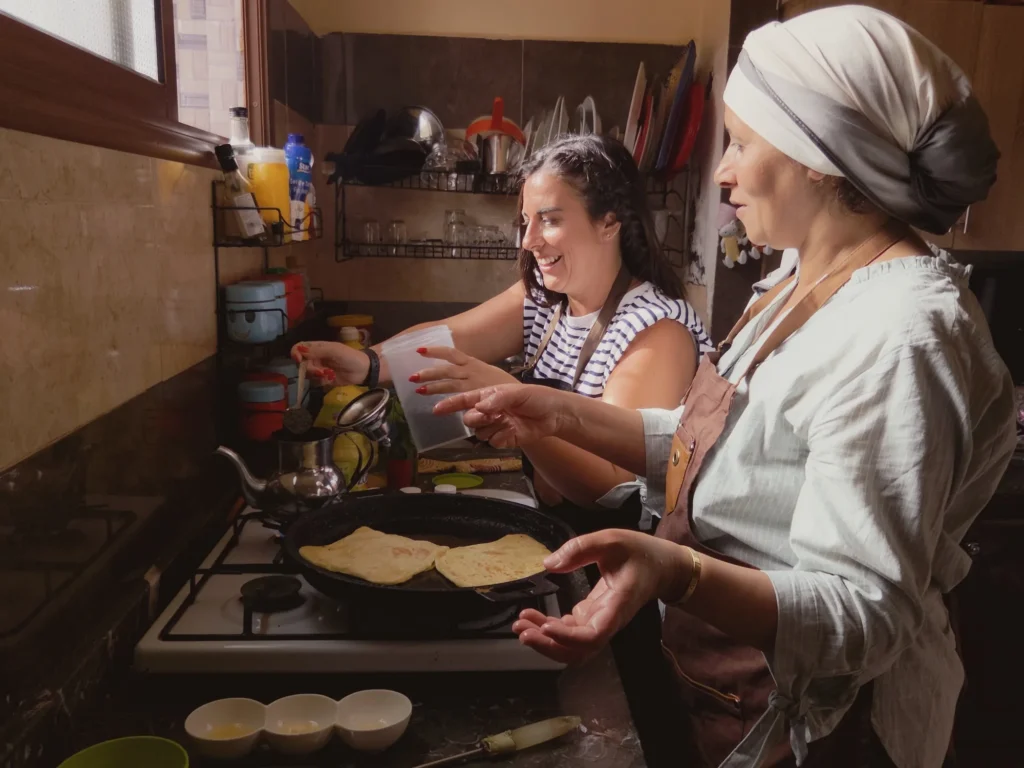 Dans sa cuisine, Aouatif cuit les msemens à la poêle avec une invitée souriante, pendant le cours msemen à Ouarzazate.