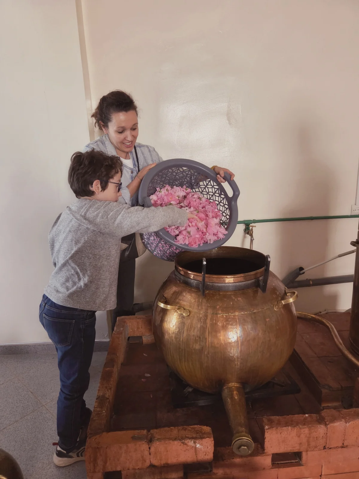Un enfant et une voyageuse versent les roses dans l’alambic en cuivre pour lancer la distillation de l’eau de rose