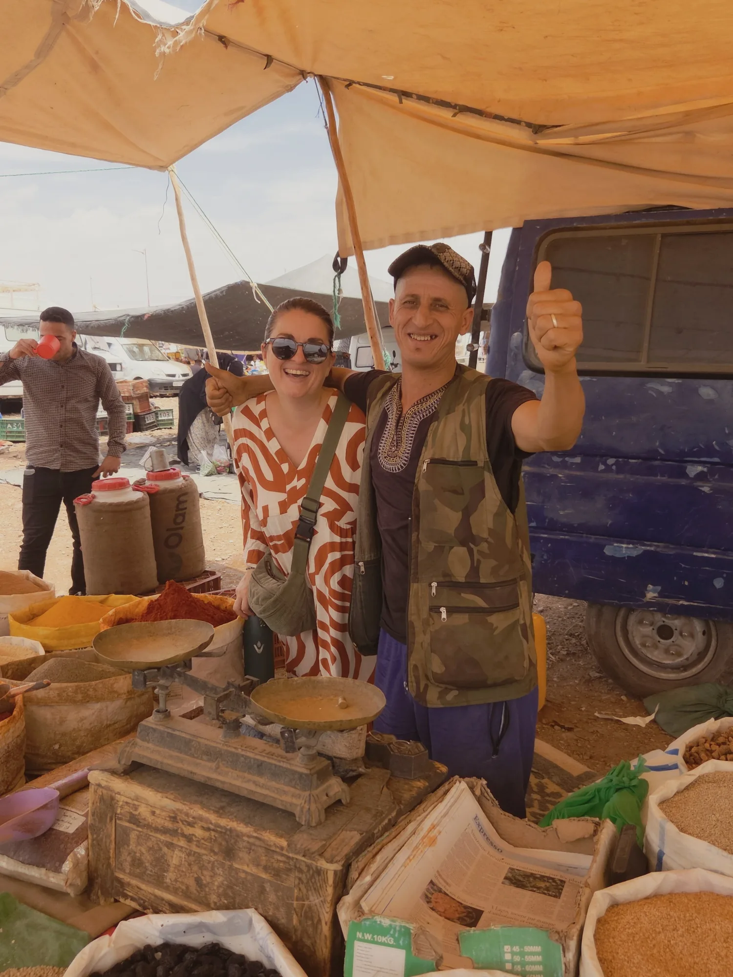 Marchand pouce levé et voyageuse souriante devant les sacs de fruits secs et d'épices à la fin du food tour au souk de Ouarzazate.