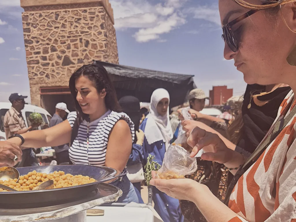 Souk de Ouarzazate : une voyageuse souriante devant l'étal de pois chiches chauds, au pied de la tour du marché