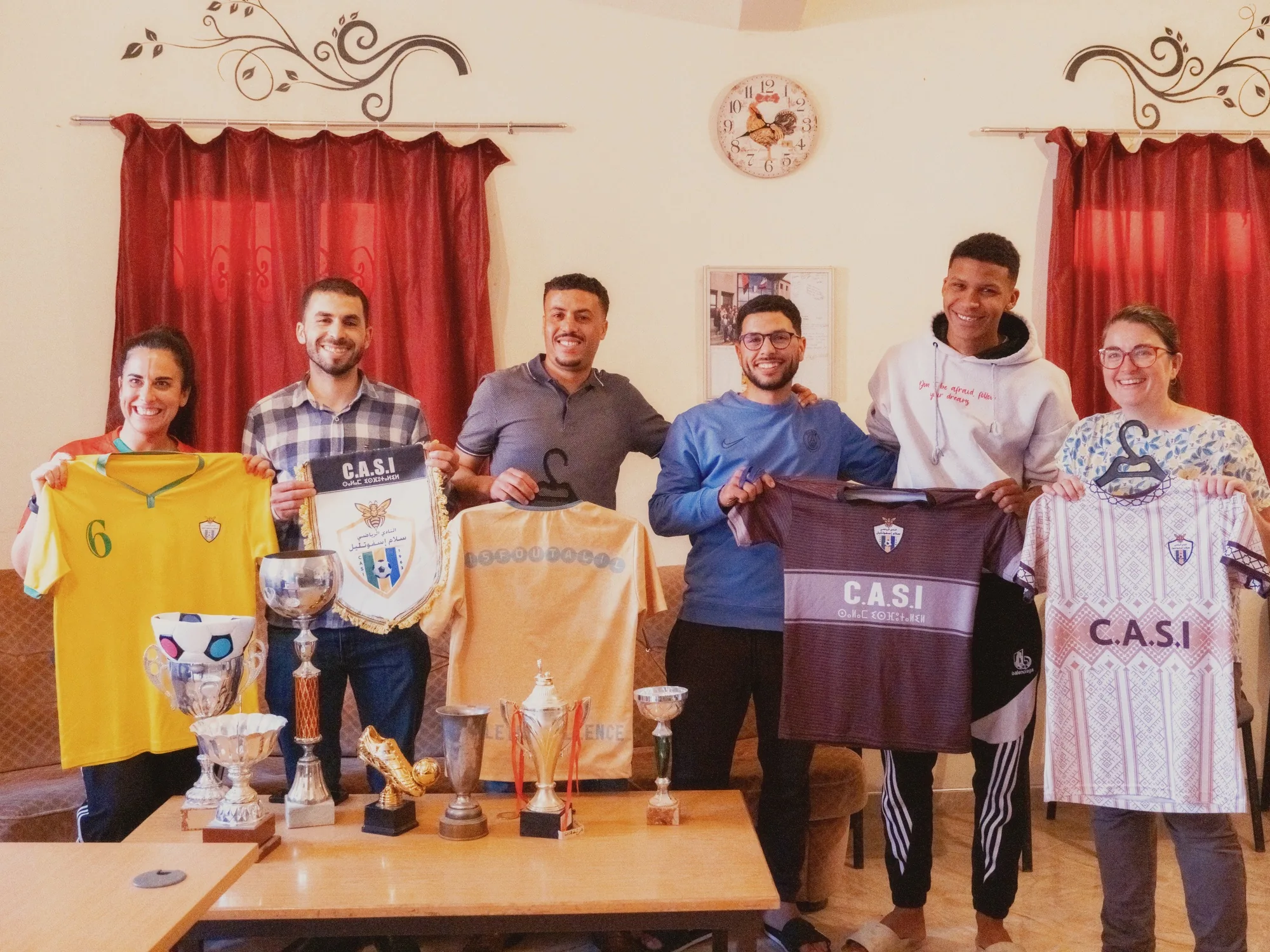 Accueil à l'association de quartier : voyageuses et entraîneurs posent avec maillots et trophées avant le foot avec les locaux à Ouarzazate.