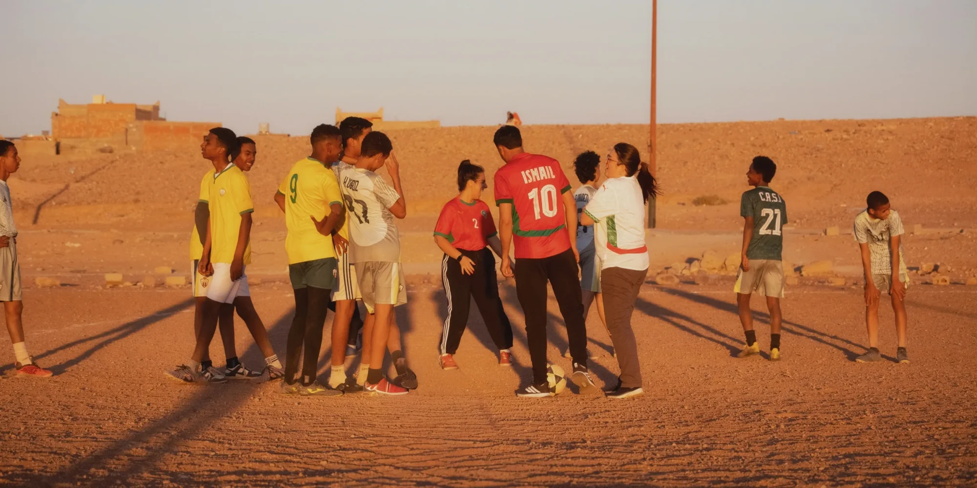 Foot avec les locaux à Ouarzazate : deux voyageuses rient avec les jeunes du quartier sur le terrain au coucher du soleil.